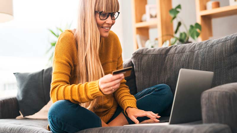 Mulher loira na casa dos 30 anos, sentada no sofá em casa, a fazer um pagamento com o cartão multibanco no portátil